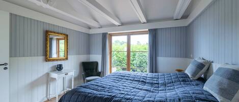 A bedroom with a large bed, a bedside table, a chair, a window with curtains, and a mirror on the wall.