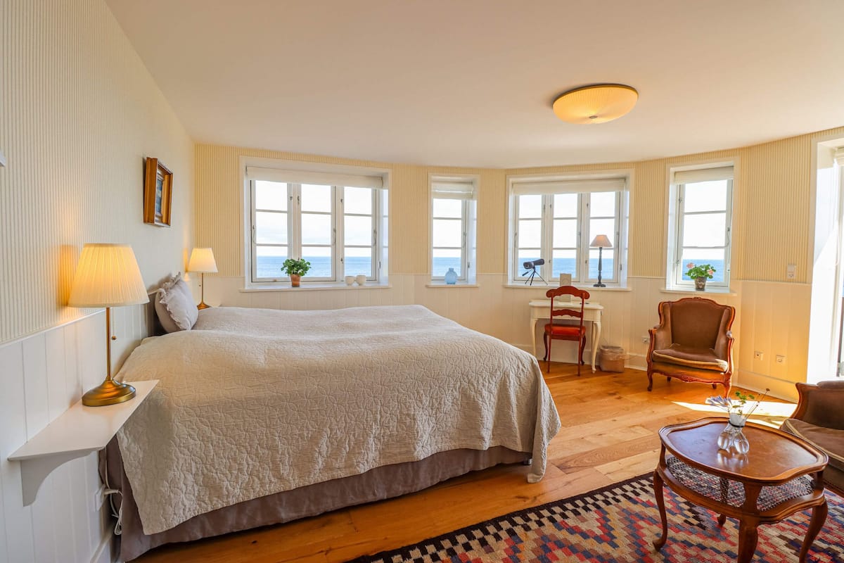 A spacious bedroom with a large bed, multiple windows, a desk, and a chair.