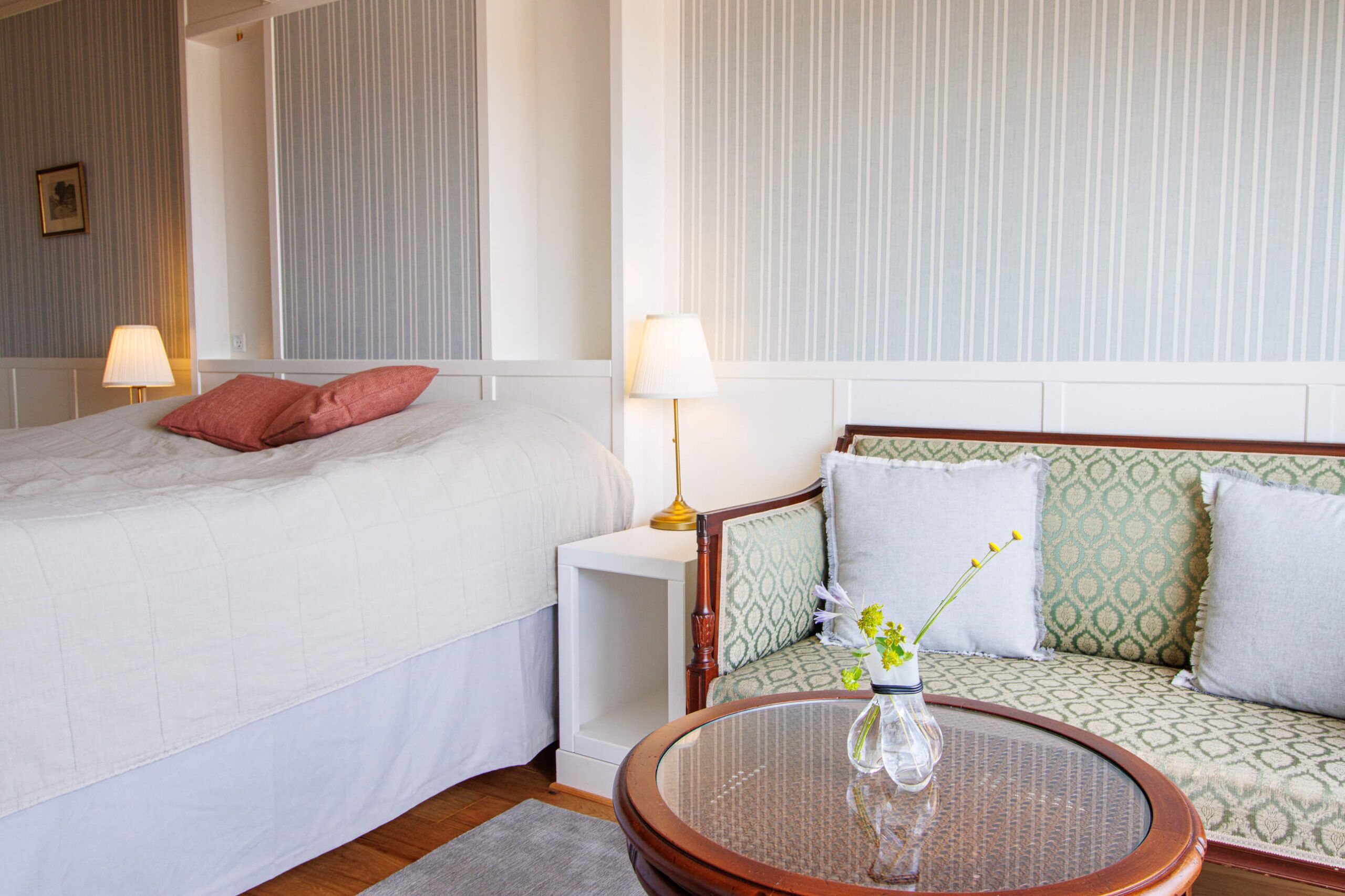 Una habitación de hotel con cama, sofá, una mesa redonda con un florero y dos lámparas.
