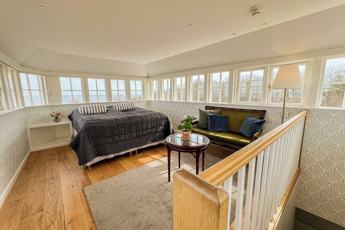 A bedroom with a bed, a sofa, a small table with a plant, and a view through large windows.