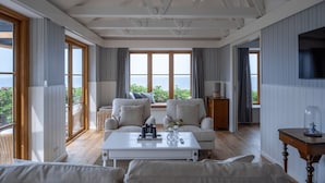 A spacious living room with a large window overlooking the sea, furnished with a white sofa, a coffee table, and a television on the wall. - Stammershalle Badehotel & Spa (Gudhjem)