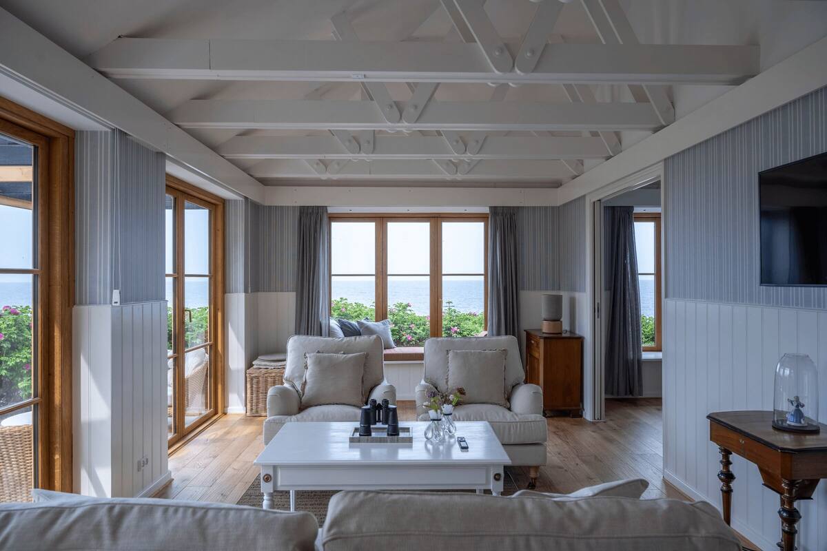 A spacious living room with a large window overlooking the sea, furnished with a white sofa, a coffee table, and a television on the wall.