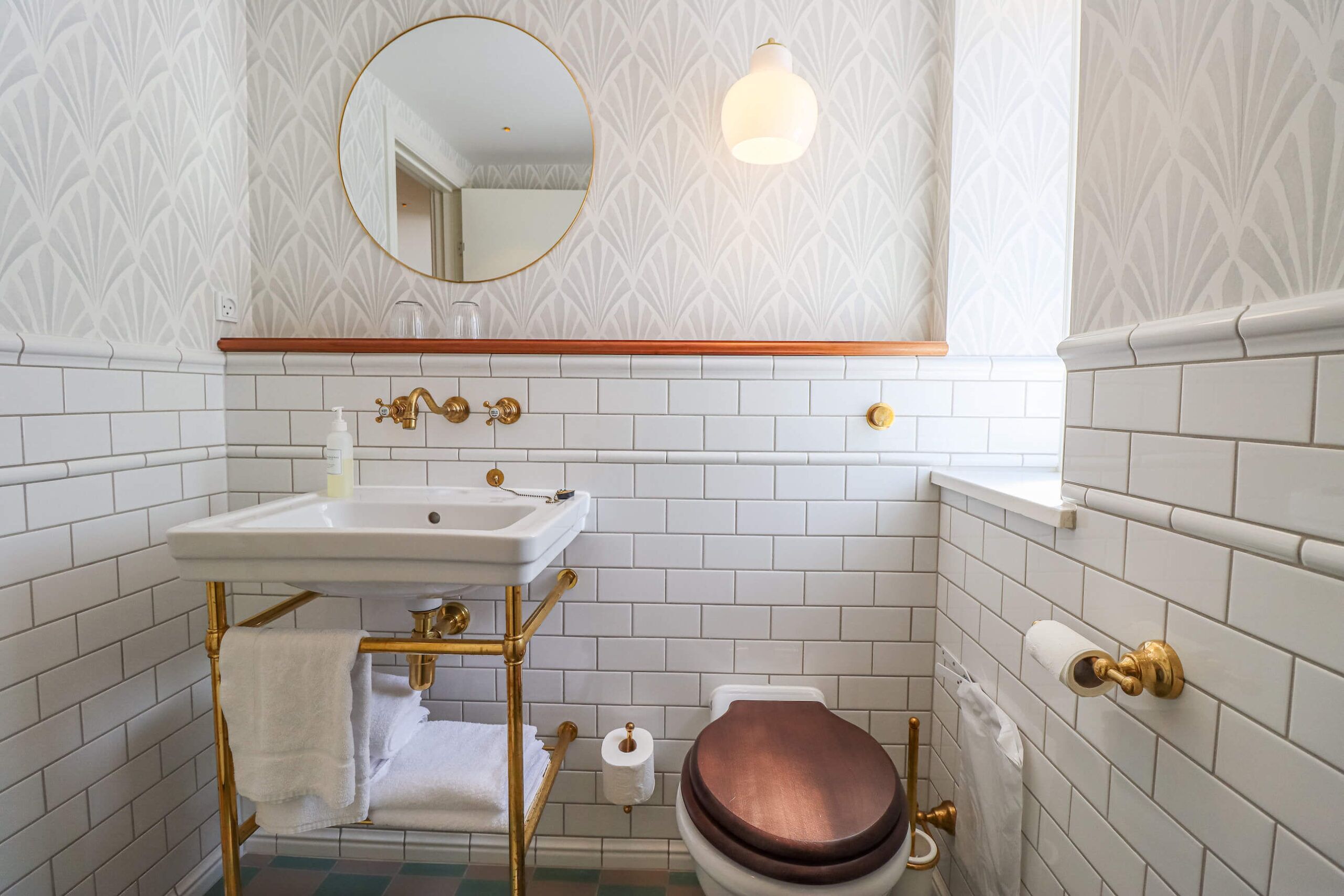 A bathroom with white subway tiles, a gold-framed sink, a round mirror, and a toilet with a wooden seat.
