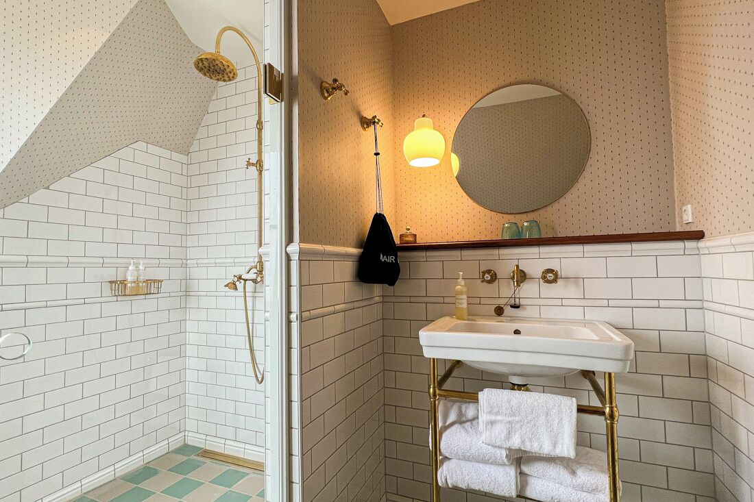 Un baño con paredes de azulejos blancos, una ducha con cabezal dorado, un lavabo con grifo dorado y un espejo sobre el lavabo.