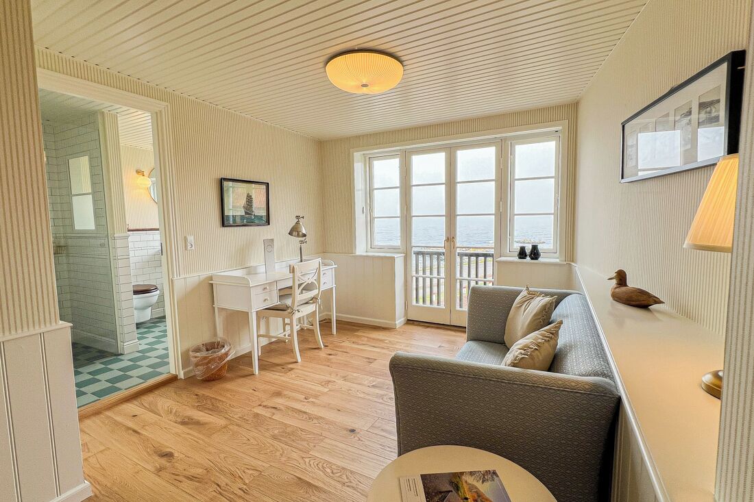 A room with a wooden floor, a grey sofa, a white desk, and a window with a view of the sea.