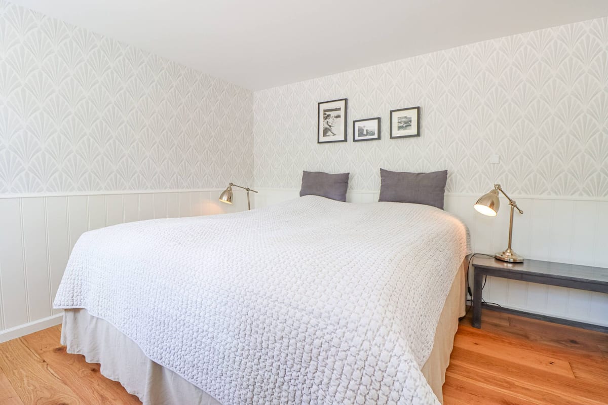 A neatly made bed with a white quilt, a bedside table with a lamp, and a patterned wall.