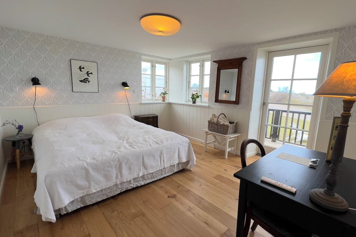 A bedroom with a large bed, two bedside lamps, a wooden desk with a lamp, a chair, and a window with a view.