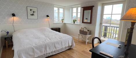 A bedroom with a large bed, two bedside lamps, a wooden desk with a lamp, a chair, and a window with a view.