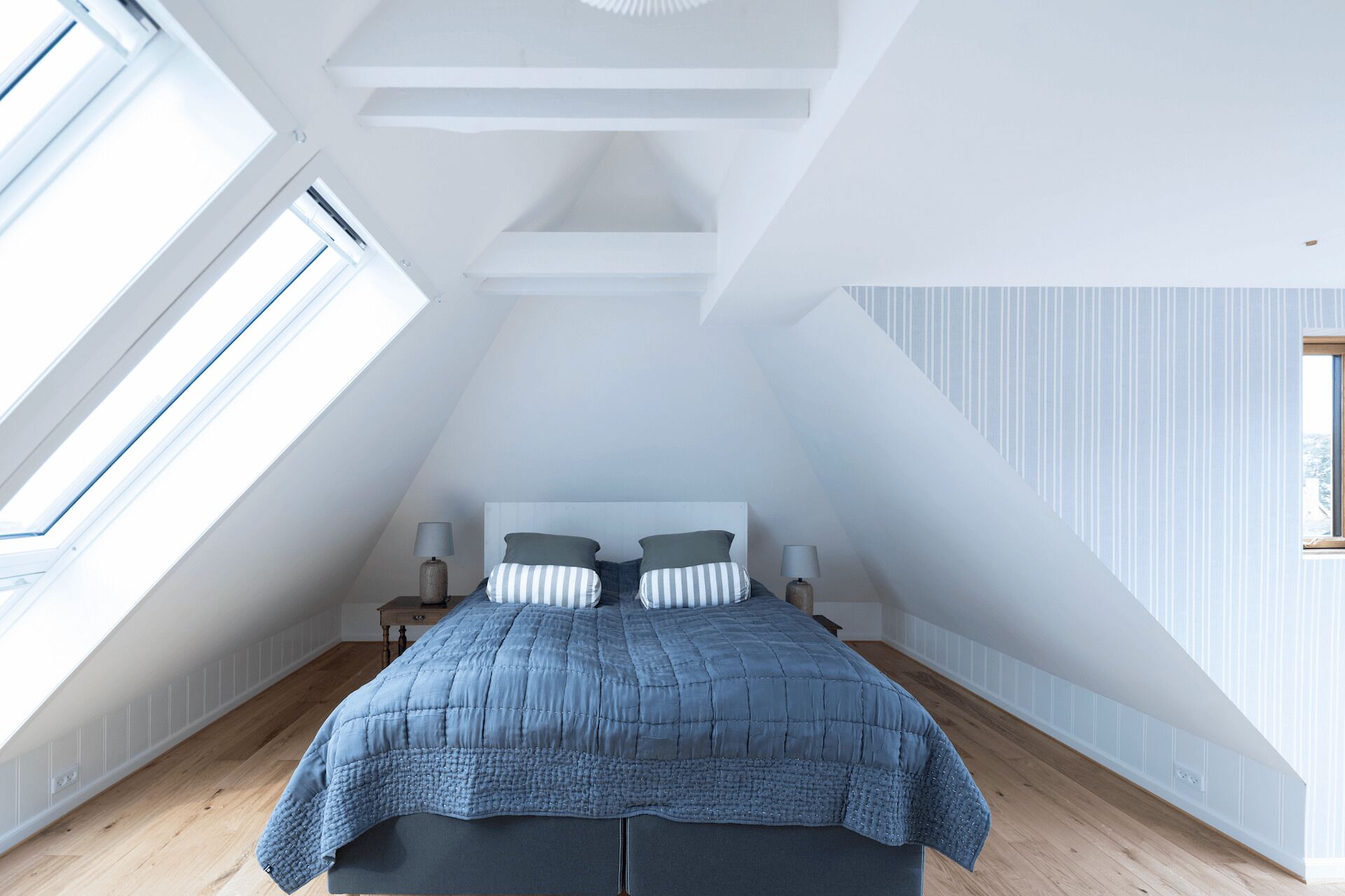 Un dormitorio con techo inclinado, una cama grande con colcha azul, dos mesitas de noche con lámparas y un ventanal que proporciona luz natural.