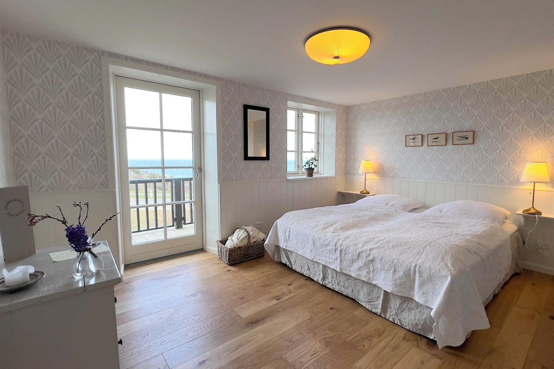 A bedroom with a large bed, two bedside tables with lamps, a window with a view of the sea, and a door leading outside.