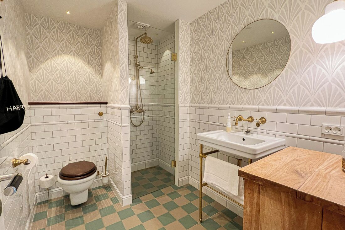 A bathroom with white tiled walls, a checkered floor, a wooden vanity, a white sink, a toilet, and a shower area.