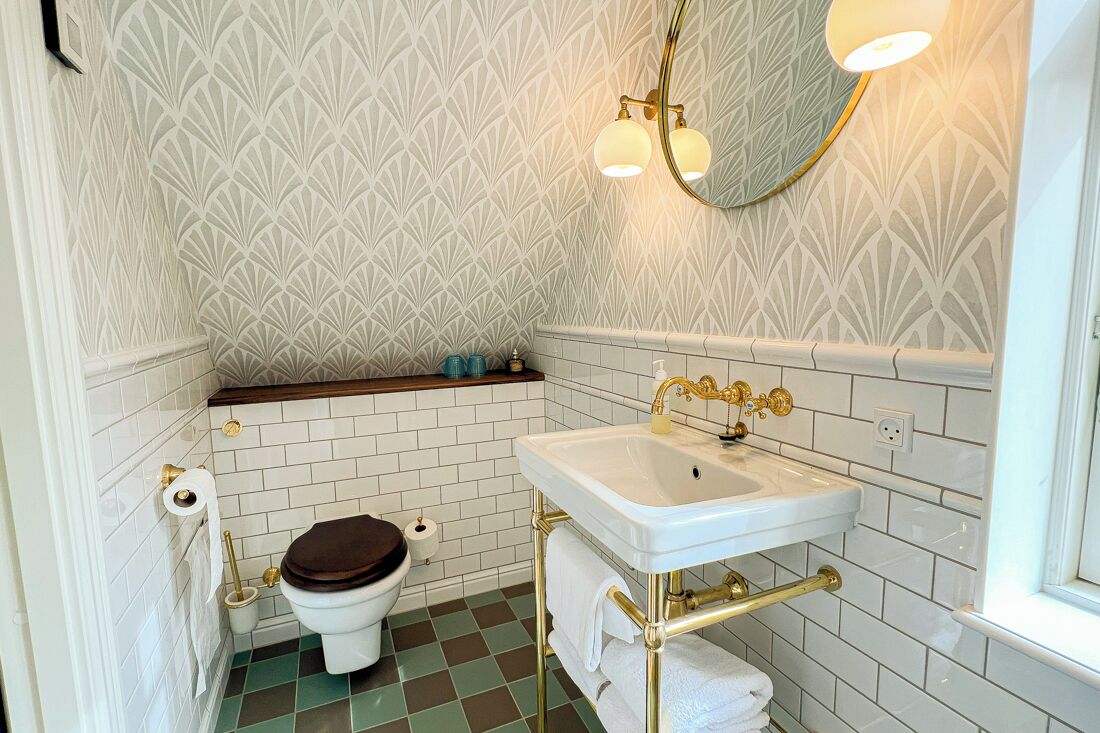 Een badkamer met een witte betegelde muur, een wit wastafelmeubel met gouden details, een toilet met een bruin zitje en een spiegel met twee lampen.