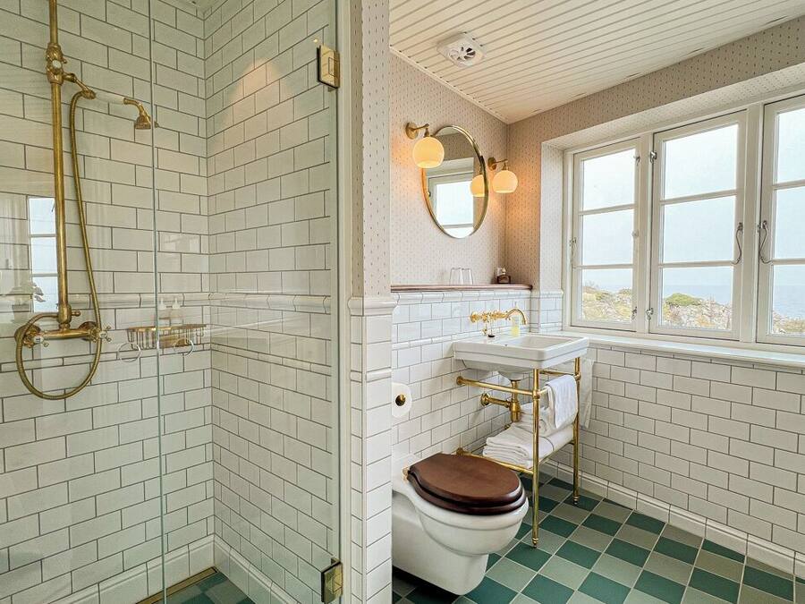 Un baño con paredes de azulejos blancos, suelo a cuadros verdes, un inodoro blanco, un lavamanos con detalles dorados y una ventana con vistas.