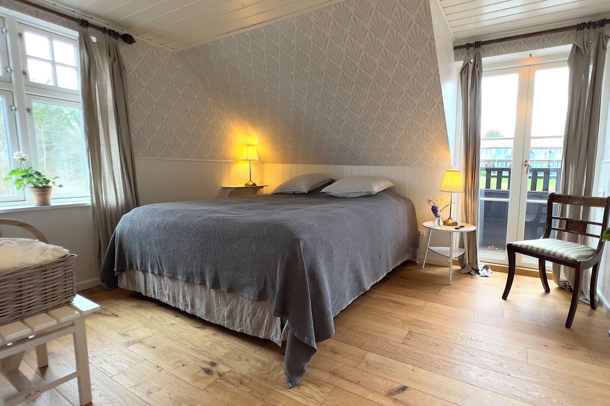 A bedroom with a large bed, two bedside lamps, a chair, a small table, and a window with curtains.