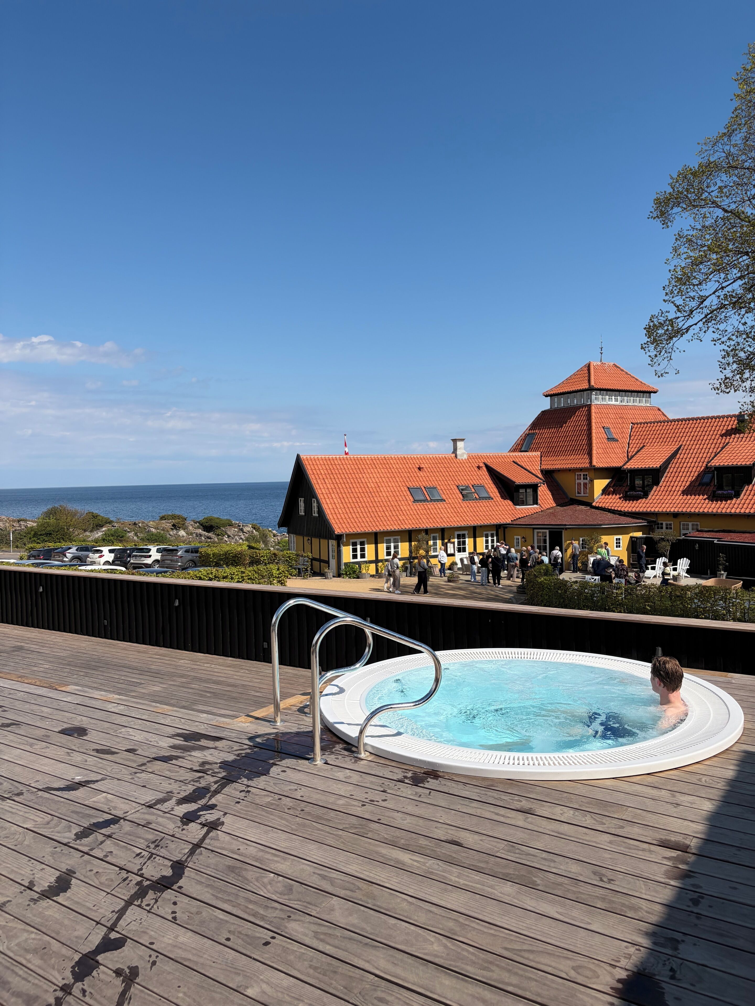 A wooden deck with a hot tub, a pool, and a building with a red roof and yellow walls.