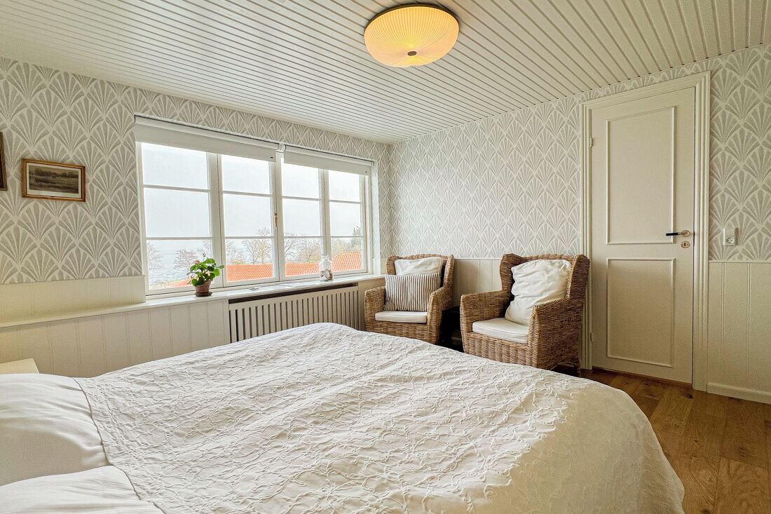 A bedroom with a large bed, two wicker chairs, a white wardrobe, and a window with a view of buildings.