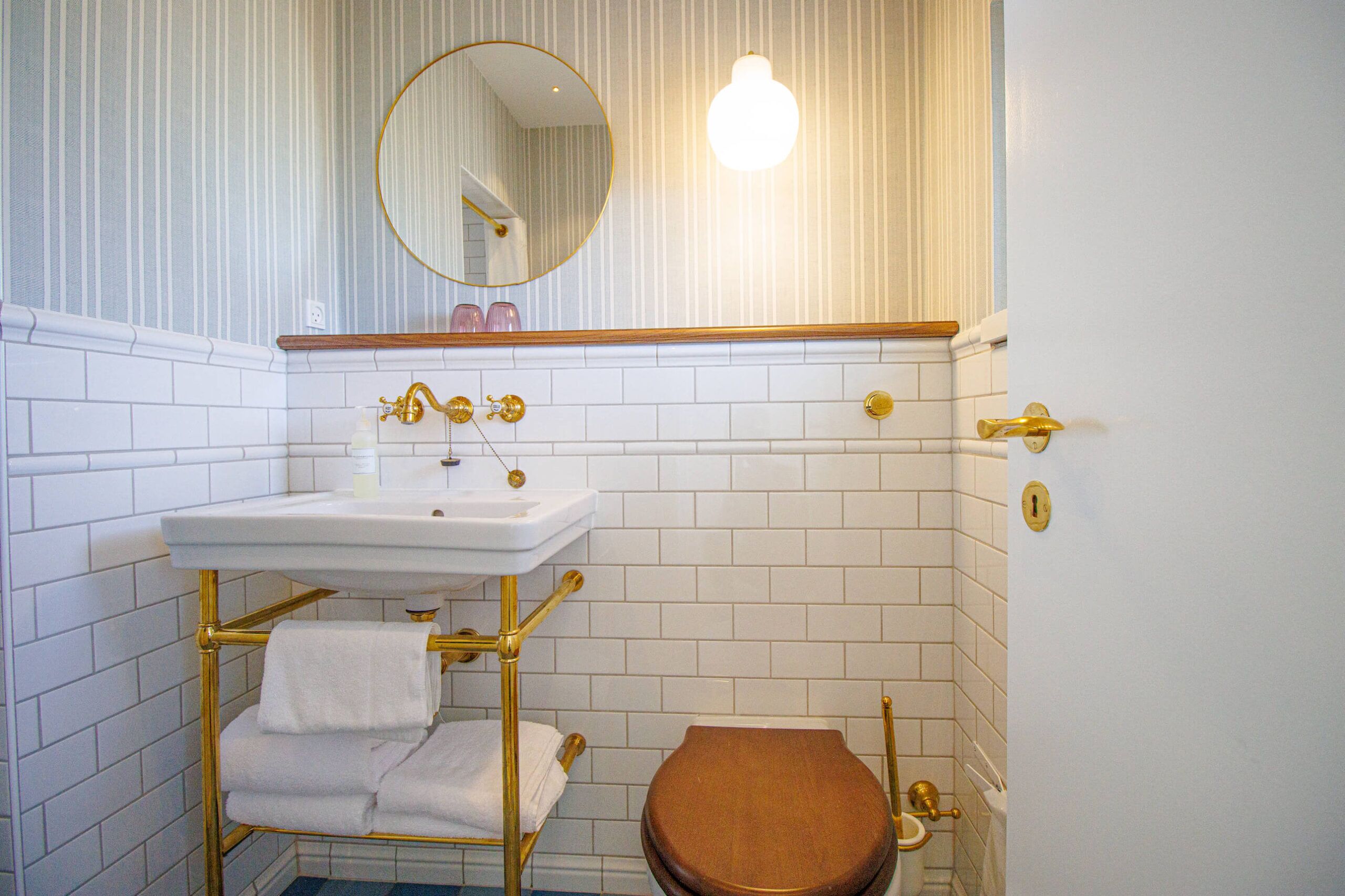 Un baño con lavamanos blanco, grifería dorada, un espejo redondo y un inodoro de madera.