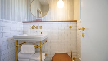 Un baño con lavamanos blanco, grifería dorada, un espejo redondo y un inodoro de madera.