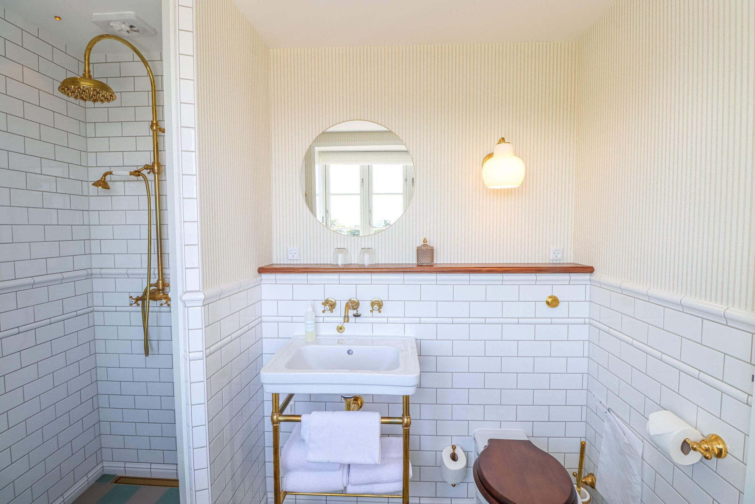 Un baño con azulejos blancos tipo metro, un cabezal de ducha dorado, un lavamanos con grifería dorada, un espejo redondo y un inodoro con palanca dorada.