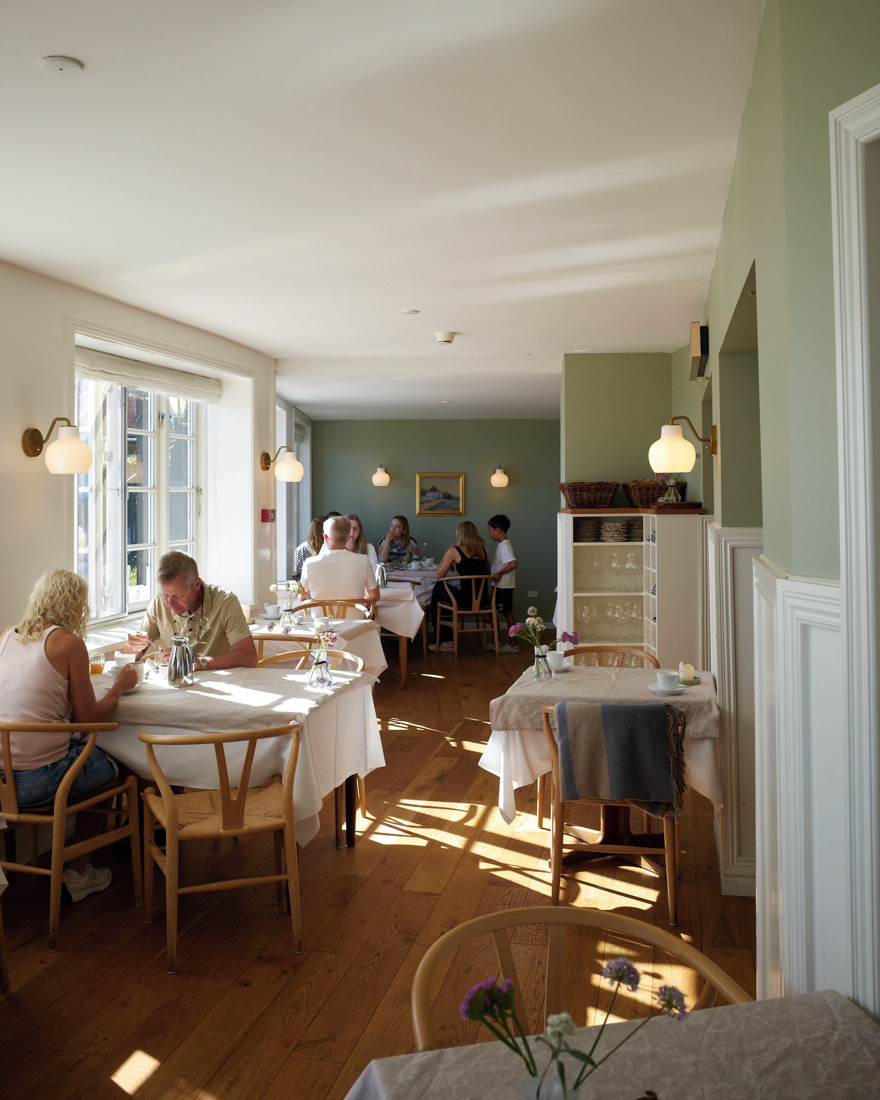 A dining area with wooden tables and chairs, a white ceiling, and a view of the street through large windows.