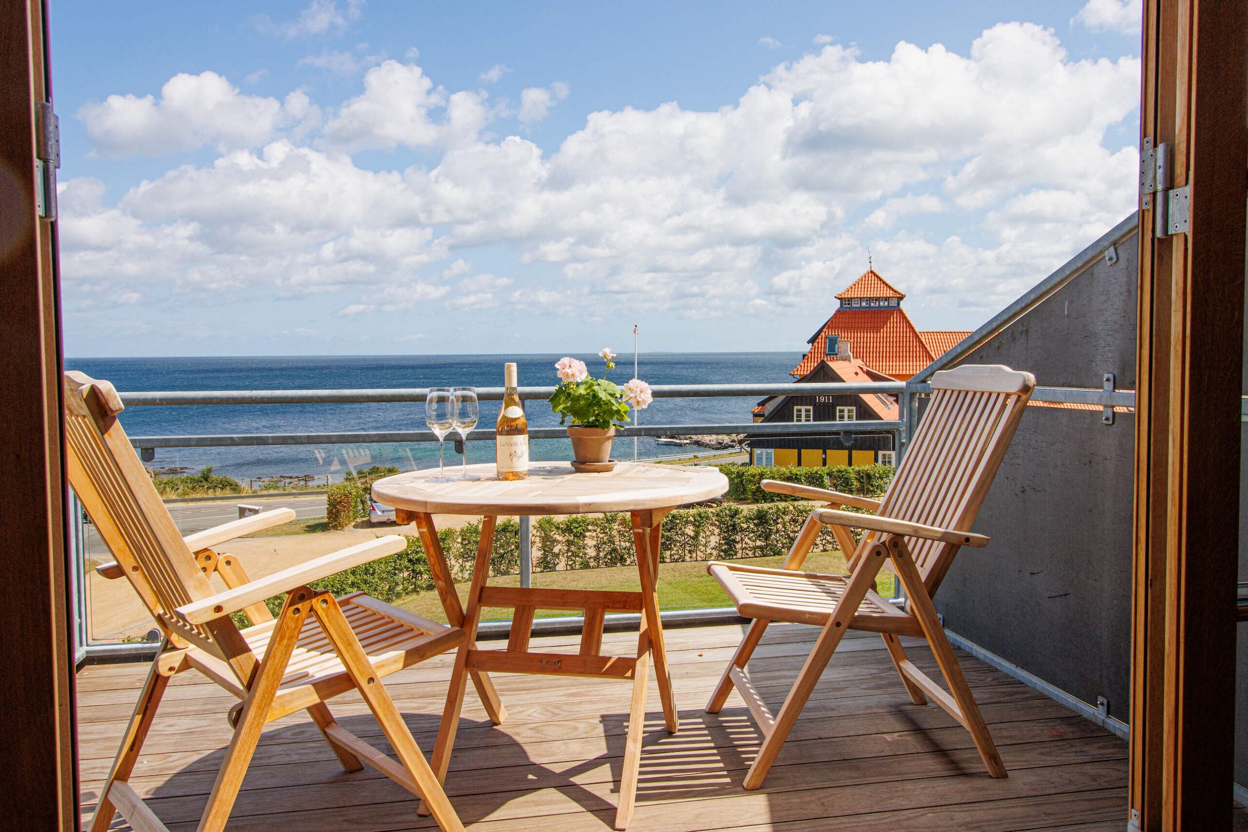 A wooden deck with two chairs and a table, overlooking a coastal view with a building in the background.