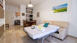 A room with a bed, a sofa, a dining table, chairs, a shelf with plants, and a painting on the wall.