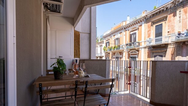 A balcony with a wooden table and chairs, a potted plant, and a view of neighboring buildings.