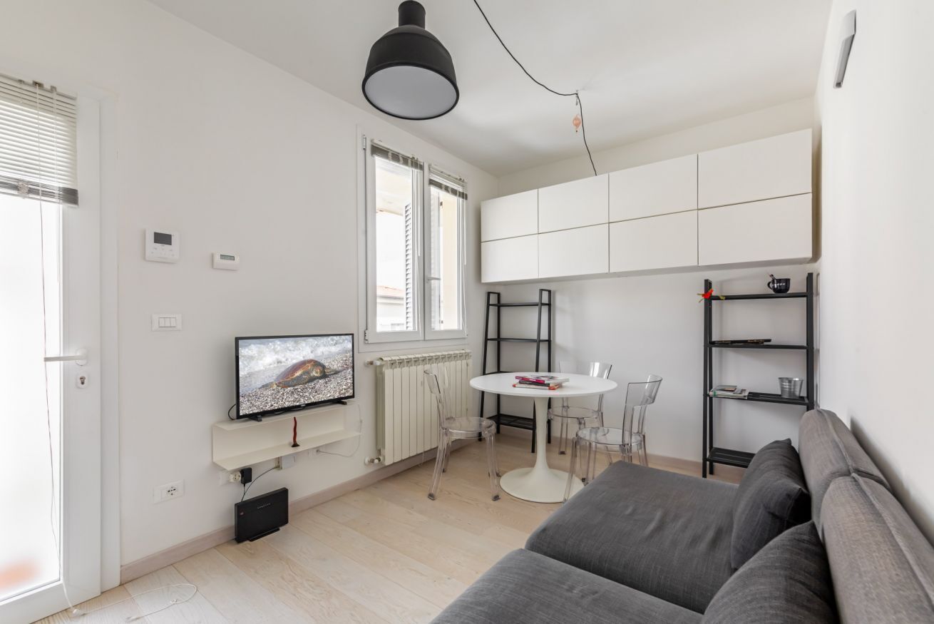 Un espacio habitable moderno con un sofá gris, una mesa de comedor blanca con sillas, un televisor y una cocina con armarios blancos.