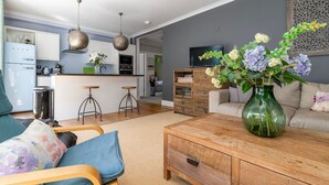 Ett modernt vardagsrum med en soffa, ett soffbord och en vas med blommor. Det finns också ett köksområde med ett kylskåp, en ugn och barstolar.