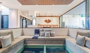 Uma sala de estar moderna com piscina integrada, área de cozinha com parede de tijolos e espaço de jantar com cadeiras de madeira.