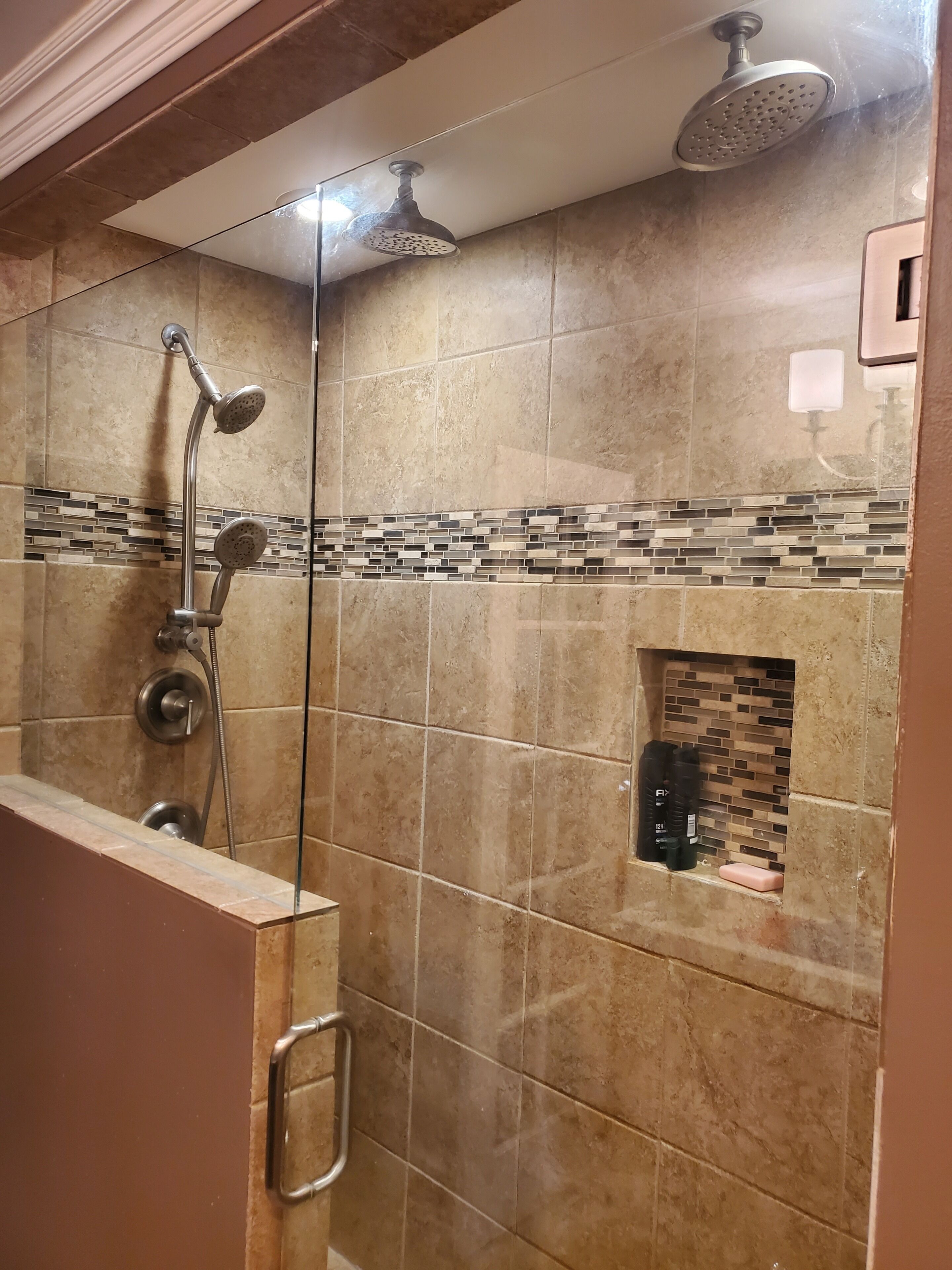 Une douche équipée d’un pommeau de douche à main et d’un pommeau de douche fixe au plafond, d’une paroi en verre et d’un mur carrelé avec une bordure en mosaïque.