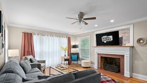 A living room with a grey sofa, a coffee table, a fireplace, a television, and a ceiling fan. - Memories on Madison | Entire Home w/ Game Room & Outdoor Fire Pit (102 Madison Ter) (Clarksville)