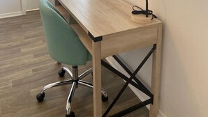 Ein kleiner, aufgeräumter Arbeitsraum mit einem Holztisch, einem grünen Bürostuhl, einer schwarzen Schreibtischlampe und einem gerahmten Zertifikat an der Wand.