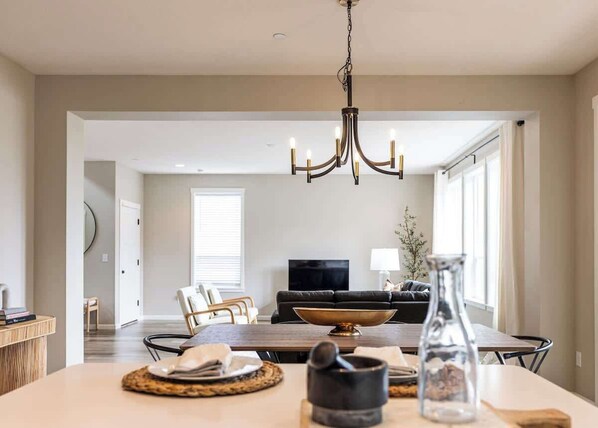 A dining area with a chandelier, a television in the background, and a dining table set with placemats and a bowl.