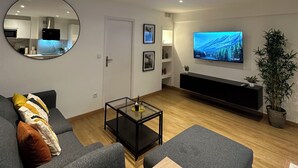 A modern living room with a grey sofa, a round mirror, a TV on the wall, and a small table. - Charmant Appart Neuf Métro Proche Confort (Bagnolet)