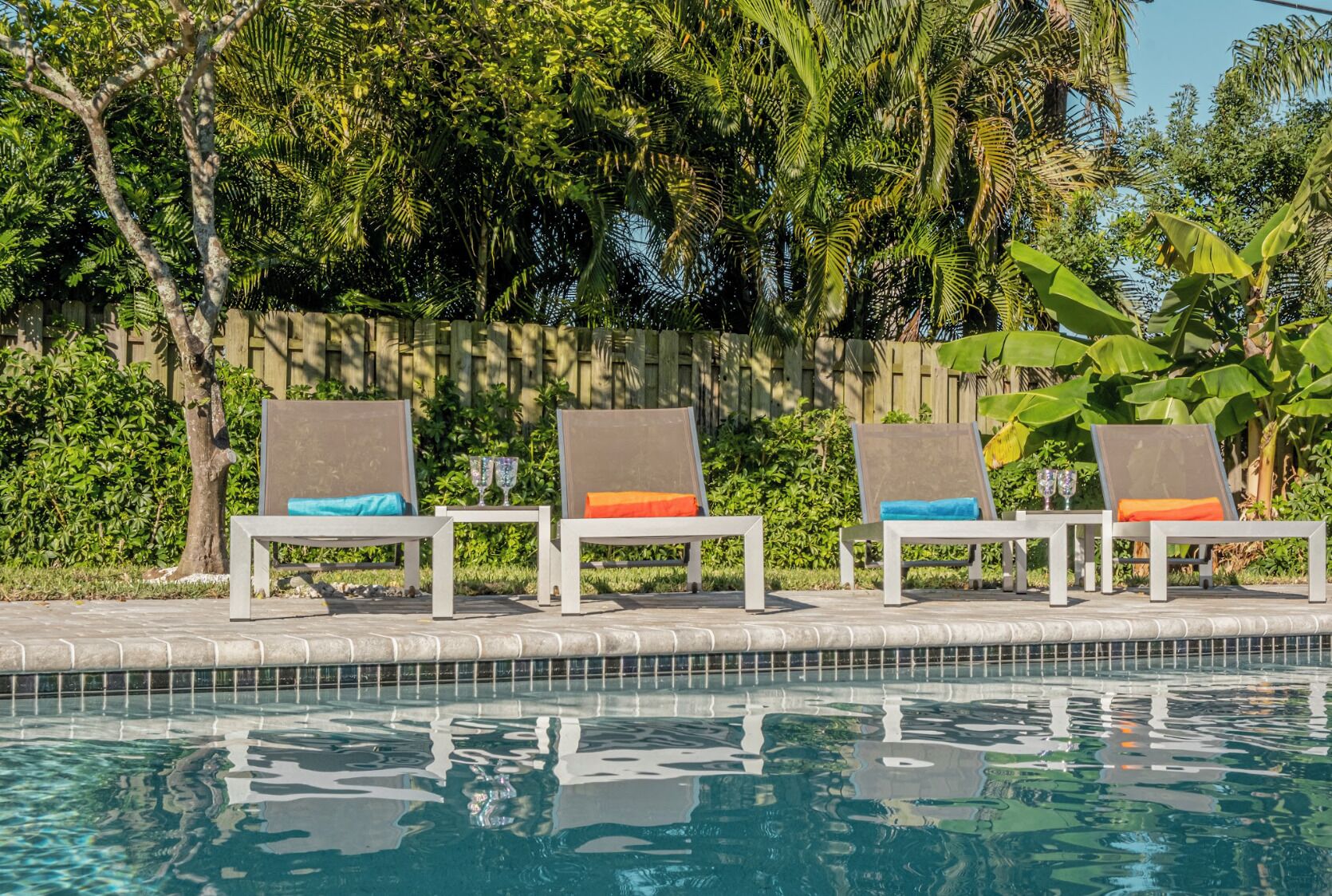 A poolside area with lounge chairs and a table, surrounded by lush greenery and a wooden fence.