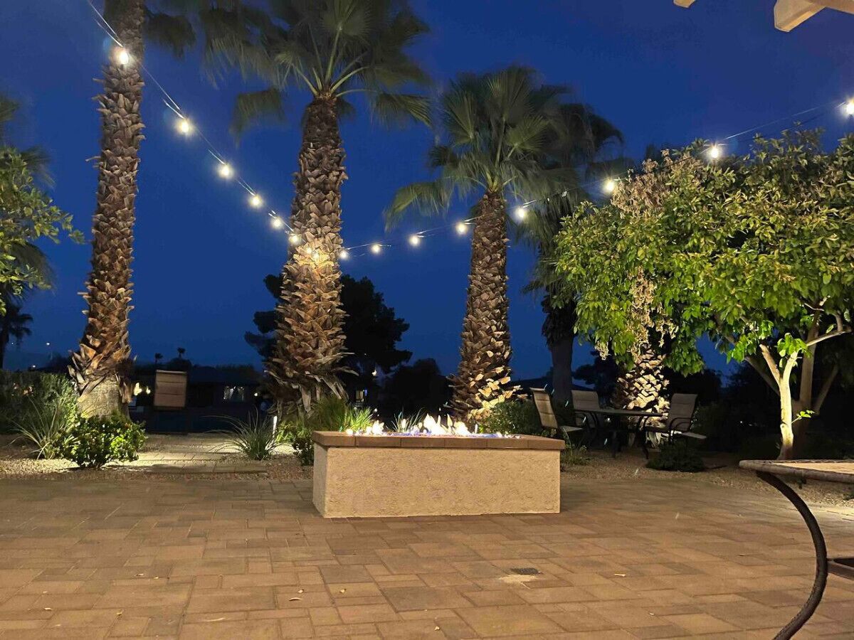 A paved outdoor area with a fire feature, palm trees, and string lights.