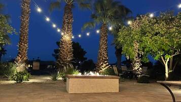 A paved outdoor area with a fire feature, palm trees, and string lights.