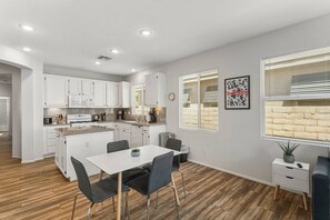 A modern kitchen with white cabinets, a central island, and a dining area with a table and chairs. - Coachella Music Festival 10min walk to grounds (Indio)