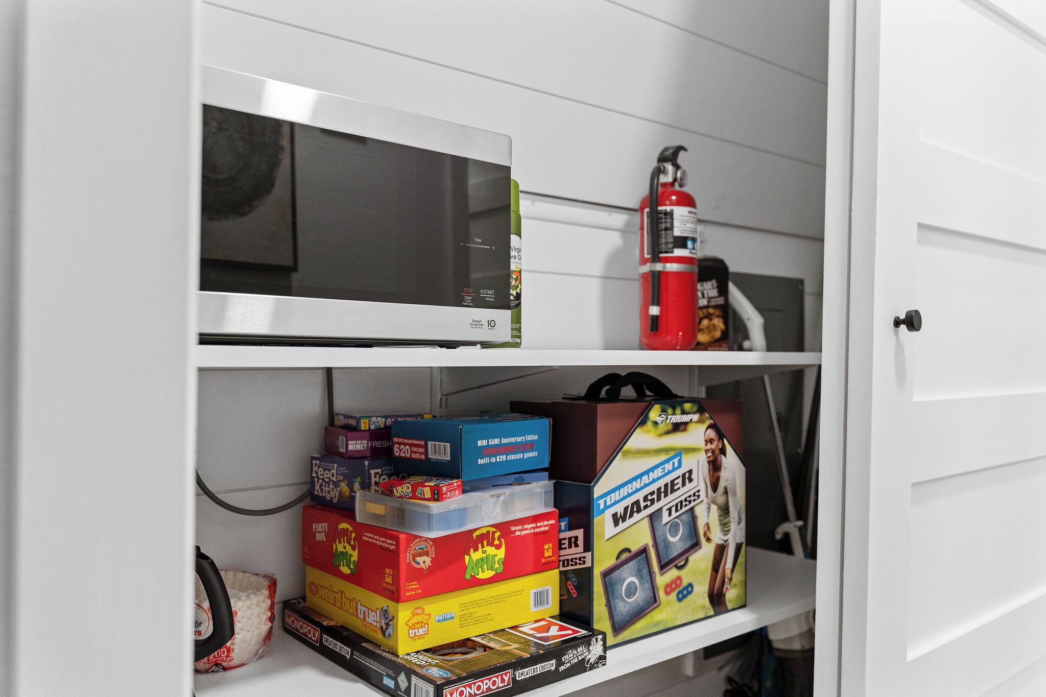 A storage cabinet with shelves containing various items including a microwave, fire extinguisher, and boxed games.