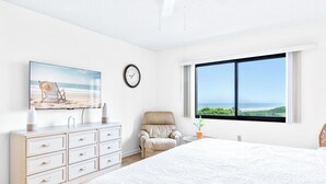 A bedroom with a large bed, a dresser, a chair, a clock, and a window with a view of the ocean. - Oceanfront Bliss | 4th Floor Condo w/ Heated Pool & Stunning Balcony Views! 🌊 (Cape Canaveral)