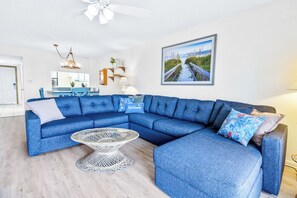 Un salon moderne avec un canapé d’angle, une table basse ronde et un tableau représentant une scène de plage accroché au mur.