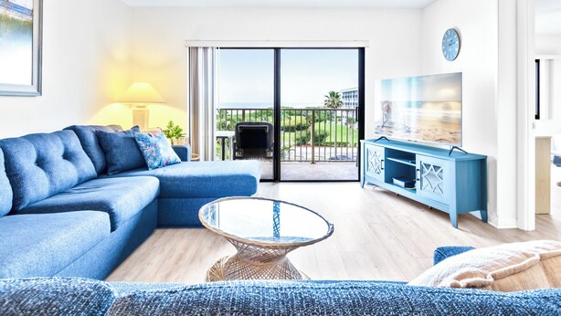 A modern living room with a blue sofa, a glass coffee table, and a TV on a blue cabinet. - Oceanfront Bliss | 4th Floor Condo w/ Heated Pool & Stunning Balcony Views! 🌊 (Cape Canaveral)