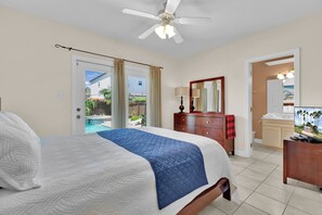 A hotel room with a bed, a dresser, a desk, a ceiling fan, and a view of the outside.