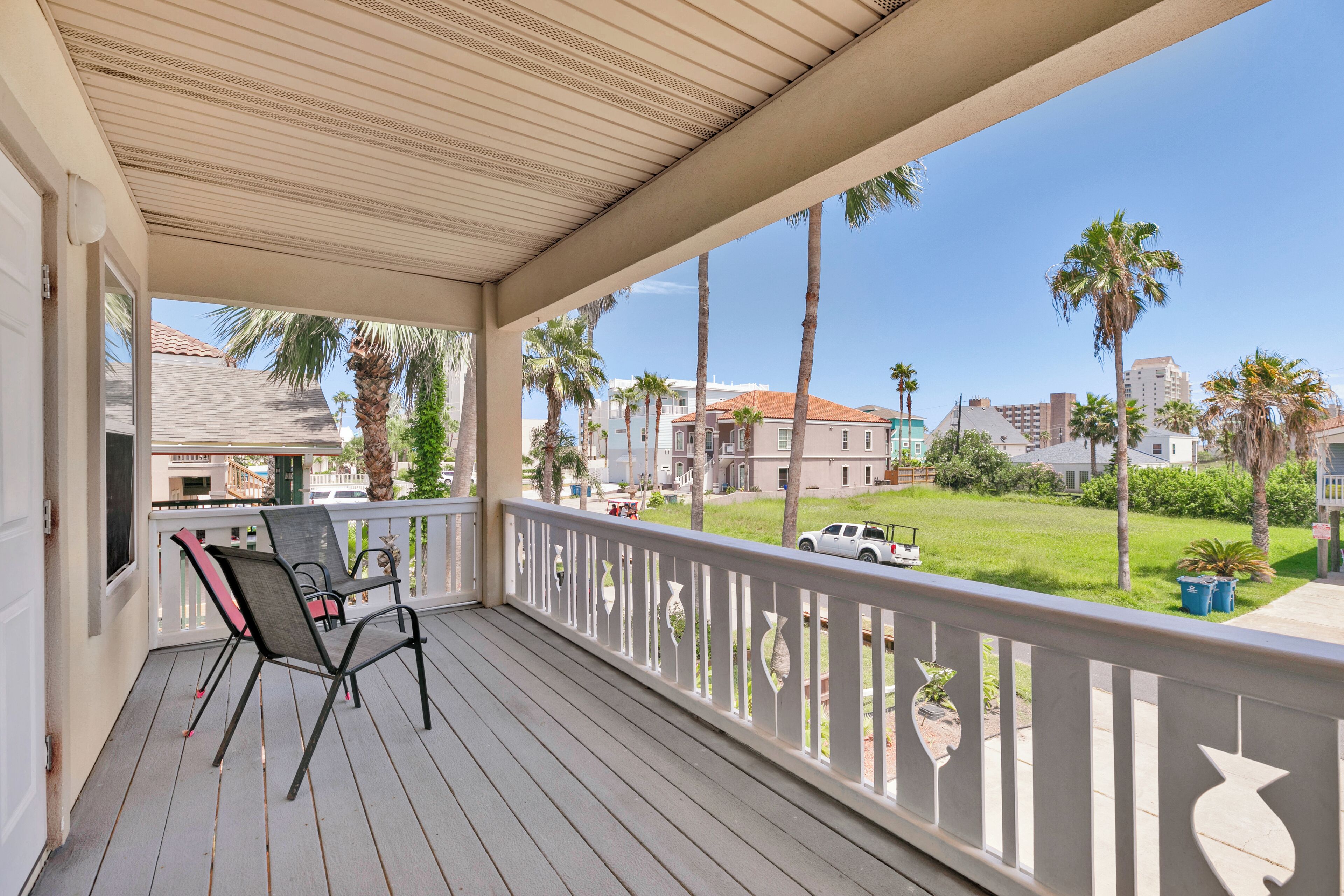 A covered porch with two chairs and a view of a residential street with palm trees and houses.