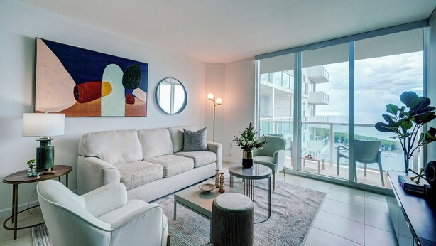 A modern living room with a sofa, armchairs, a coffee table, and a large window offering a view of a building and a body of water. - Direct Bayview One Bedroom Condo in Coconut Grove-includes parking (Coconut Grove)