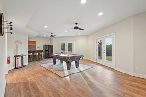 Amplia habitación con mesa de billar, ventiladores de techo y vistas a través de amplias puertas correderas de vidrio.
