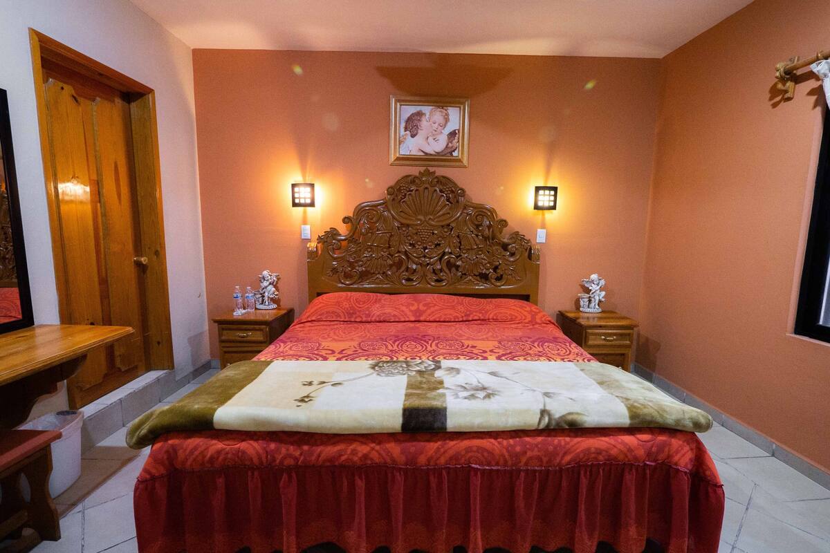 A bedroom with a wooden headboard, two bedside tables with lamps, a bed with a red bedspread, and a wall-mounted picture.