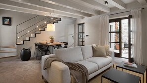 Una sala de estar moderna con un sofá, una mesa de centro y un área de comedor con una mesa y sillas. Hay una escalera con barandilla, una puerta que conduce a otra habitación y una ventana con cortinas.