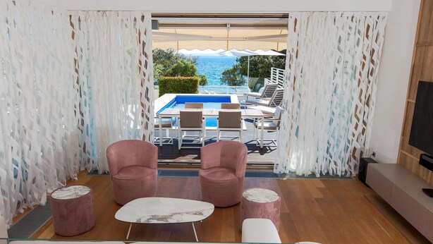 A modern living room with a glass coffee table, two pink armchairs, and a sofa. A pool area with a table and chairs is visible through the curtains. - Beach front home in Petrcane (Petrcane)